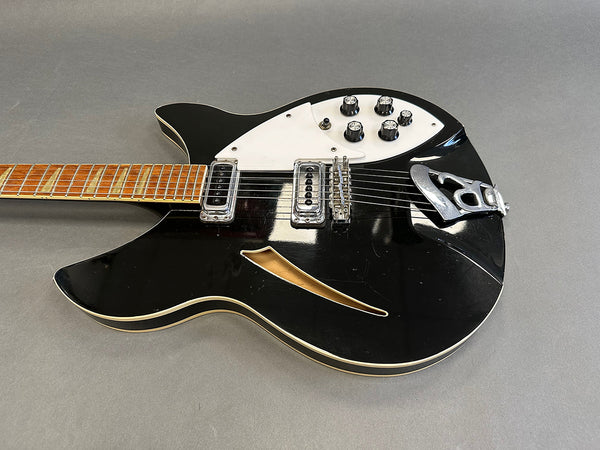 Black semi-hollow body electric guitar with white pickguard, six knobs, two humbucker pickups, and a Bigsby-style tailpiece on gray background