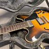 Semi-hollow body electric guitar with sunburst finish lying in a plush gray guitar case with black handle, showing detailed neck, pickups, and control knobs on a gray surface.