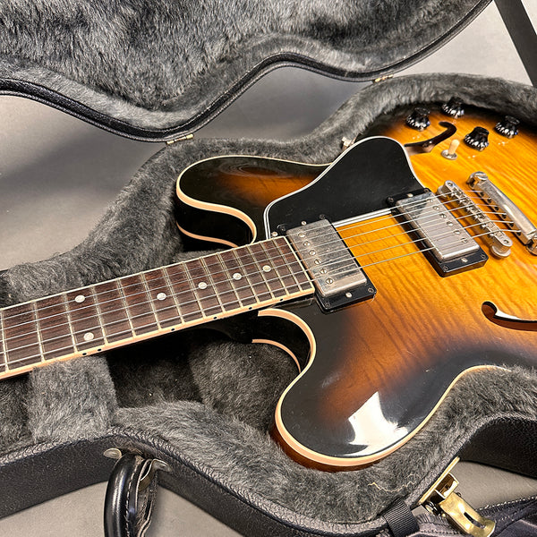 Semi-hollow body electric guitar with sunburst finish lying in a plush gray guitar case with black handle, showing detailed neck, pickups, and control knobs on a gray surface.