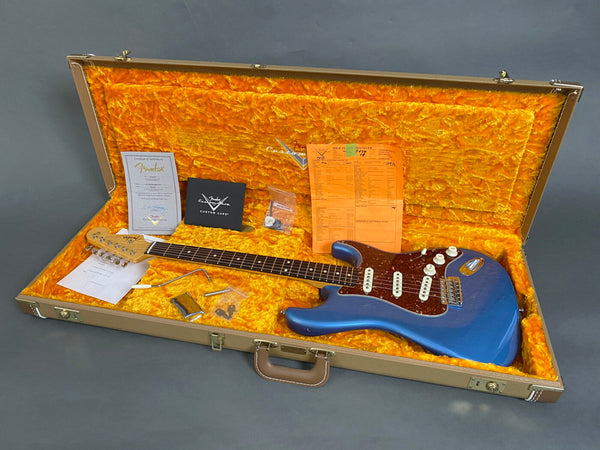 Fender Custom Shop electric guitar in blue with tortoiseshell pickguard inside a tan hardshell case with orange plush interior, accessories, and certificate of authenticity arranged around the guitar.