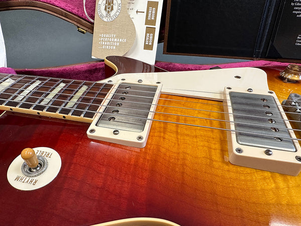 Close-up of Gibson Les Paul electric guitar body showing sunburst finish, humbucker pickups, toggle switch labeled Rhythm and Treble, and part of the guitar neck with inlays.