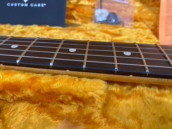 Close-up of electric guitar fretboard and strings with dot inlays, resting on plush yellow guitar case lining, showing maple neck and custom care guitar accessories in the background.