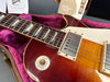 Close-up of Gibson Les Paul electric guitar neck and body with sunburst finish, visible pickups, triangular fretboard inlays, and rhythm-treble switch in hard case with purple lining and Gibson Custom paperwork.