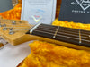 Close-up of Fender Custom Shop guitar neck and headstock showing strings, frets, and tuning pegs with certificate and case interior in background