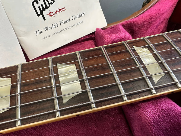 Close-up of Gibson guitar rosewood fretboard with mother-of-pearl block inlays and metal frets inside a pink plush-lined guitar case showing Gibson Custom booklet
