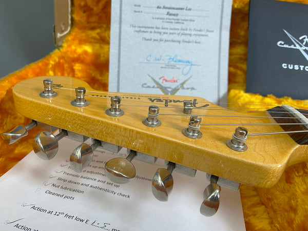Close-up of Fender Stratocaster electric guitar headstock with vintage tuning pegs and strings, displayed in orange plush-lined guitar case with Fender Custom Shop certificate and inspection checklist in background
