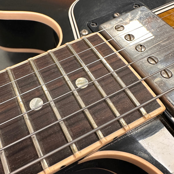Close-up of electric guitar neck showing rosewood fretboard, metal frets, pearl dot inlays, and vintage metal humbucker pickup on black guitar body