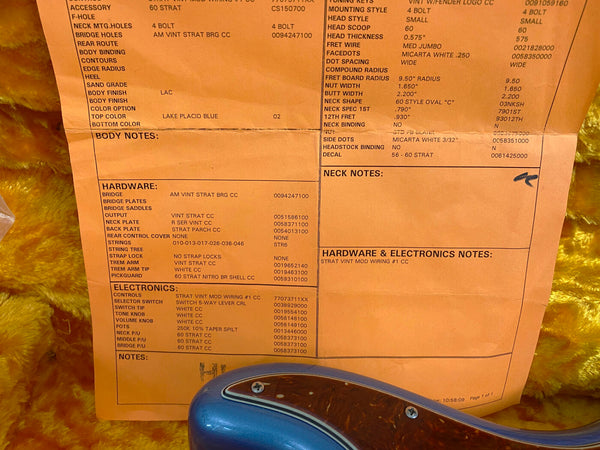 Vintage Stratocaster guitar specification sheet with hardware and electronics details, showing Lake Placid Blue top color and 60 Strat pickups, placed on a yellow textured fabric background with a partial view of the guitar body.