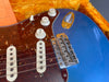 Close-up of blue electric guitar body with tortoiseshell pickguard, three white single-coil pickups, tone and volume control knobs, and chrome bridge inside a yellow plush lined case