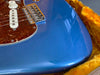 Close-up of blue electric guitar body with tortoiseshell pickguard, white volume knob, and metal bridge inside yellow plush guitar case showing fine finish cracks and edge wear.