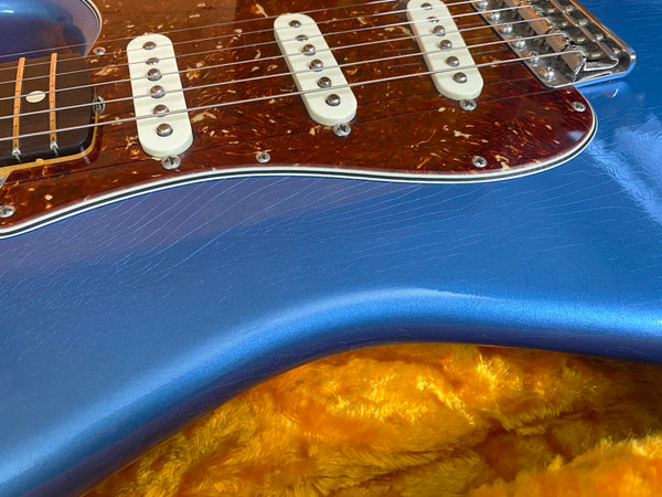 Close-up of blue electric guitar body with tortoiseshell pickguard and white single-coil pickups on orange plush guitar case interior