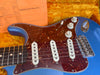 Close-up of blue electric guitar with tortoiseshell pickguard, three single-coil pickups, tone and volume knobs, and chrome bridge on yellow plush case background with orange spec sheet