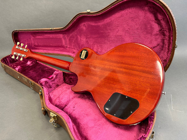 Back side of a sunburst Les Paul style electric guitar resting in an open brown hard shell case with magenta velvet interior lining