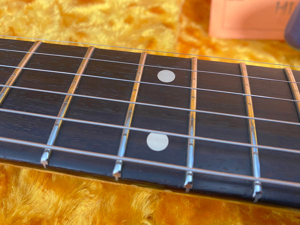 Close-up of guitar fretboard with metal frets and dot inlays on dark wood, steel strings visible, yellow guitar case lining background