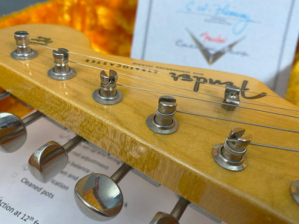 Close-up of Fender Stratocaster electric guitar headstock with tuning pegs and strings, natural wood finish, and Fender logo visible.