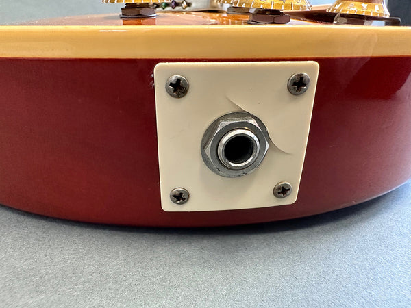 Close-up of vintage electric guitar output jack plate with visible crack on cream-colored plastic mounting plate and red guitar body.