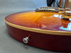 Close-up of sunburst electric guitar body showing bridge, control knobs, and strap button on reddish-brown finish with cream binding.