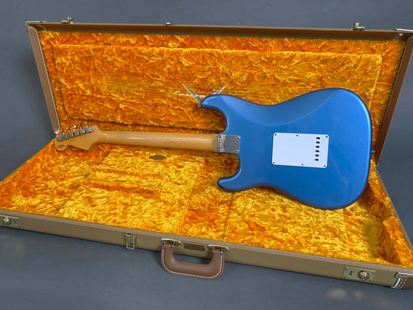Back view of blue Fender electric guitar with maple neck inside open tan vintage-style hard case lined with plush orange interior