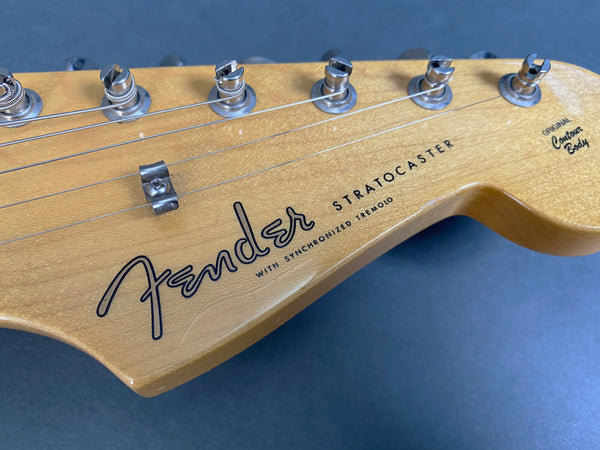 Close-up of light wood Fender Stratocaster guitar headstock with tuning pegs and strings, featuring "Original Contour Body" and "with Synchronized Tremolo" text on a dark background