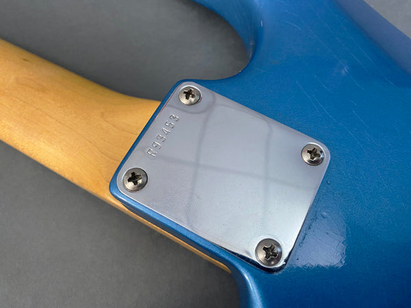 Close-up of blue electric guitar neck plate with serial number R93453 and four screws, maple neck and metallic blue body finish with light scratches.