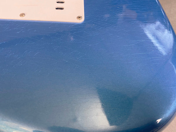 Close-up of glossy blue electric guitar body showing fine crackle finish texture and white plastic backplate with screws