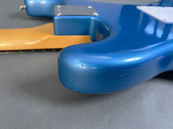 Close-up of blue electric guitar body with visible scratches and dents, showing neck joint and natural wood neck on gray background.