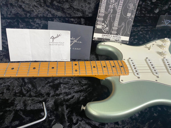 Close-up of Fender Stratocaster electric guitar neck and body in metallic seafoam green finish with white pickguard, included owner's manual, Fender 5-position pickup selector switch instructions, and tremolo arm resting on black plush guitar case interior.