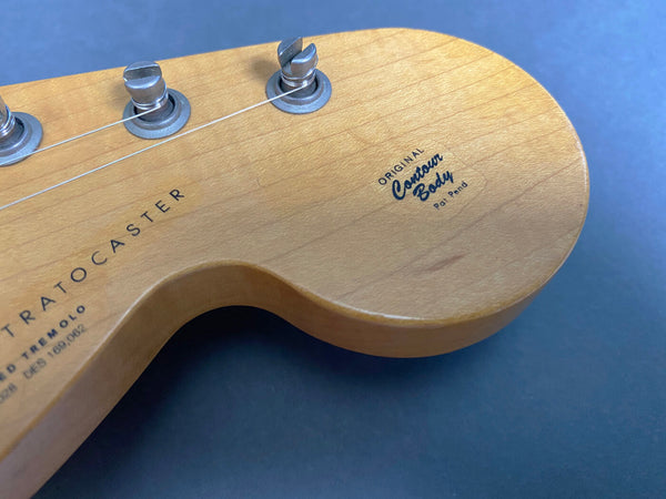 Close-up of natural wood guitar headstock with vintage tuning pegs and Original Contour Body logo on a Strato Caster electric guitar against a blue background