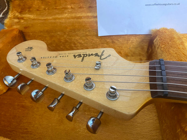 Close-up of Fender Stratocaster guitar headstock with tuning pegs and strings in an orange plush-lined guitar case, showing maple wood finish and rosewood fretboard, featuring the Fender logo and model details.