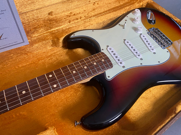 Electric guitar with sunburst finish and white pickguard displayed inside plush orange guitar case with certification paper in corner