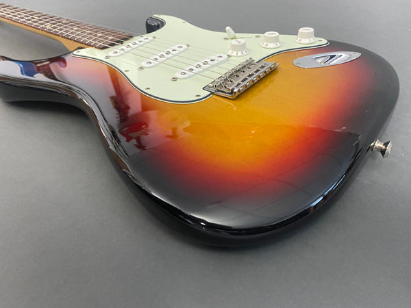 Sunburst electric guitar with white pickguard, three single-coil pickups, tone and volume knobs, and chrome bridge on gray background.