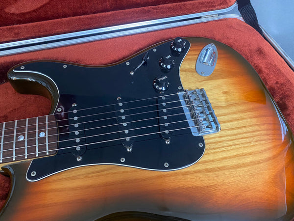 Electric guitar with sunburst finish, black pickguard, three single-coil pickups, volume and tone control knobs, shown in red velvet-lined guitar case