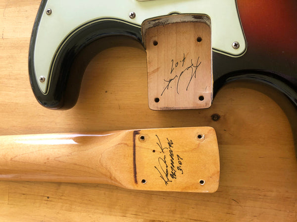 Close-up of detached guitar neck and body joint showing handwritten signatures and date on natural wood finish, part of a vintage electric guitar with sunburst finish and white pickguard on wooden surface.