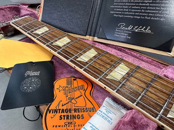Close-up of a Gibson Custom Shop guitar neck with mother-of-pearl block inlays, displayed inside a pink plush-lined guitar case with Gibson Vintage Reissue Strings and a Gibson Custom tag.