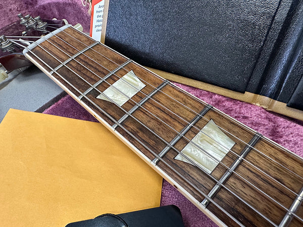 Close-up of guitar neck with mother-of-pearl block inlays on rosewood fretboard, metal frets, and strings, next to a yellow envelope and guitar case lining