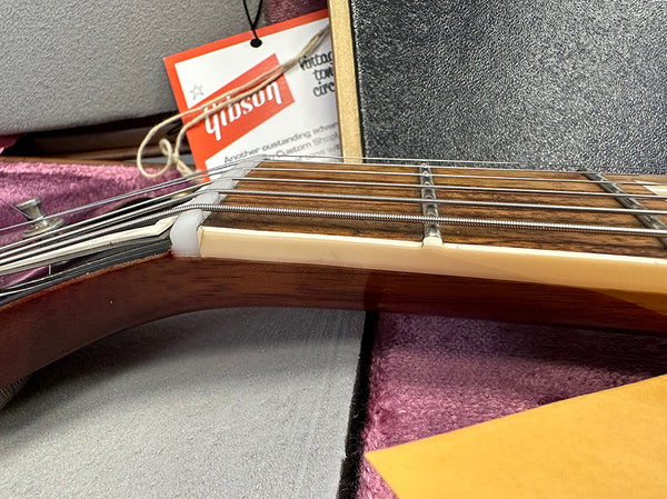 Close-up of guitar neck and nut with six metal strings, showcasing rosewood fretboard and cream binding, with a Gibson tag visible in the background.