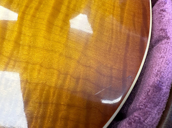 Close-up of honey burst guitar body with visible scratch and wear inside purple plush-lined guitar case