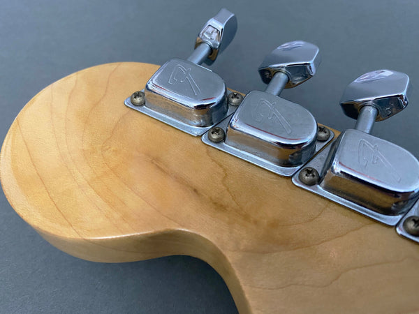 Close-up of maple guitar headstock with vintage chrome tuning pegs featuring engraved logos on a gray background