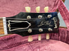 Close-up of black Gibson Les Paul Model guitar headstock with six tuning pegs, white nut, and strings on purple plush guitar case lining