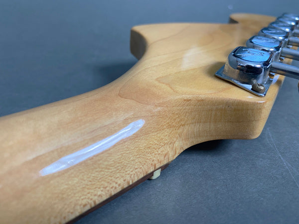 Close-up of a guitar neck headstock showing natural wood finish with chrome tuning pegs and a visible white scratch or mark on the wood.