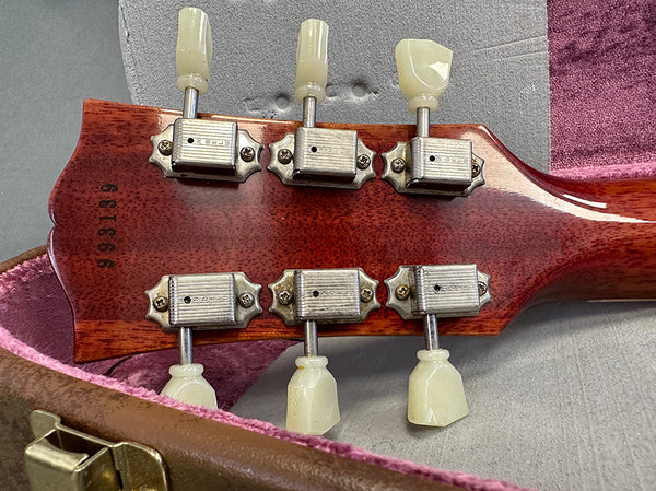 Close-up of vintage guitar back headstock showing six metal tuning pegs with cream plastic buttons and serial number stamped on wood inside an open guitar case
