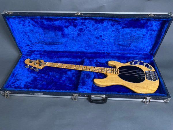 Natural wood finish electric bass guitar with black pickguard in blue velvet-lined hard case on gray background