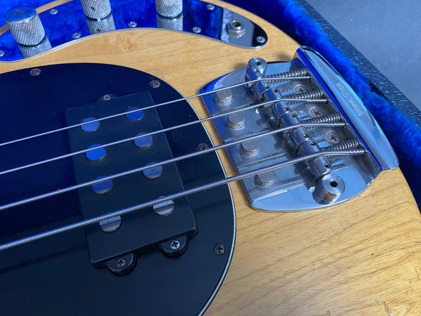 Close-up of electric bass guitar bridge, pickups, and control knobs on natural wood finish body with blue trim case interior