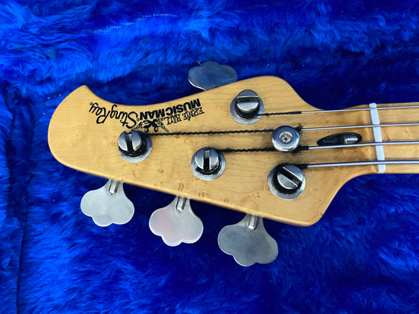 Maple headstock of Purple Bull Music Man Stingray bass guitar with tuning pegs on blue plush background