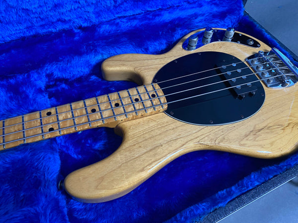 Natural wood finish electric bass guitar with black pickguard and maple neck, displayed on blue plush guitar case interior