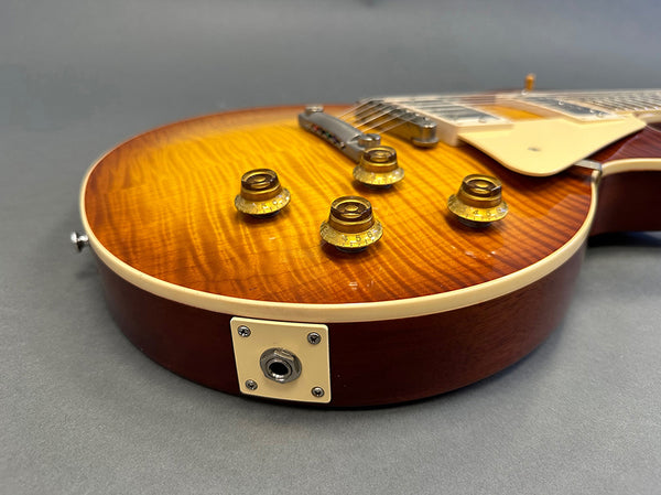 Close-up of sunburst electric guitar body with amber control knobs and chrome bridge, showing input jack and quilted maple top finish on gray background