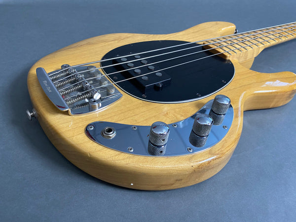 Natural wood finish electric bass guitar body with black pickguard, chrome bridge, and control knobs on gray background