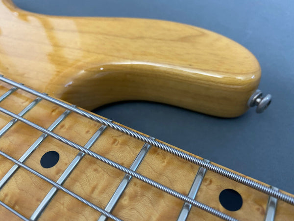 Close-up detail of natural wood grain bass guitar neck with metal frets and round black fret markers on gray background