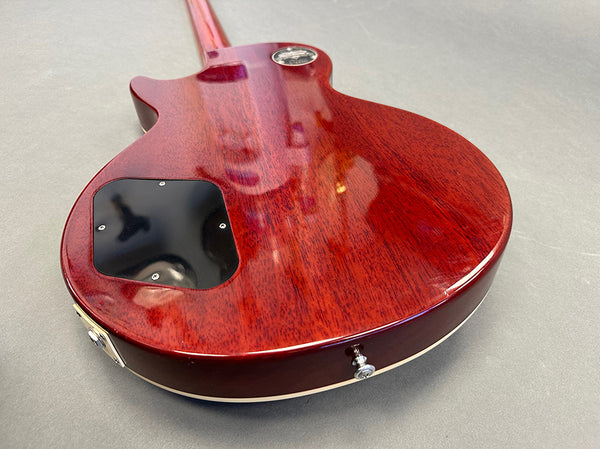 Back view of red mahogany electric guitar body with black control cavity cover and strap button on gray background