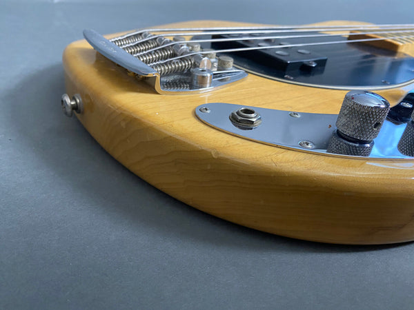 Close-up of natural finish electric bass guitar body showing chrome bridge, control knobs, and input jack on gray background